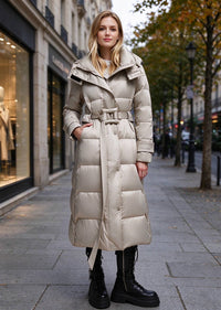 A woman stands on a city sidewalk wearing a long, light beige puffer coat with a belted waist and large collar, paired with black lace-up boots. The background shows a blurred urban street scene with storefronts and trees.