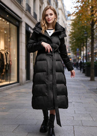 A woman wearing a long black puffer coat with a belted waist and high collar, standing on a city sidewalk.