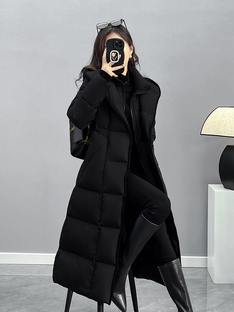 A woman poses in a long, unzipped black down coat, revealing a monochromatic black outfit underneath. She has sunglasses perched on her head and is wearing knee-high black leather boots.