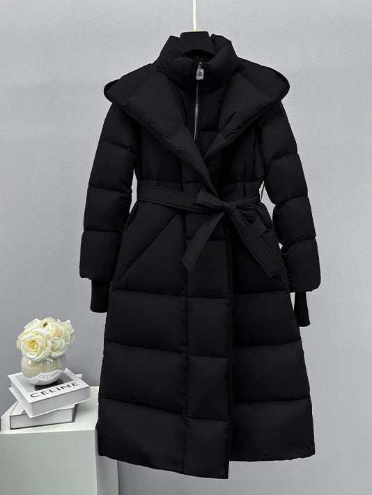 A front-view studio shot of a long black puffer coat hanging on a hanger. The coat features a high collar, a wrap-style front secured with a matching fabric belt, and ribbed knit cuffs.