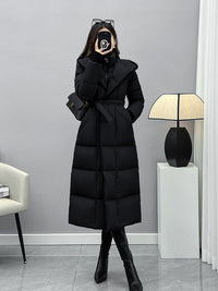 A full-length mirror selfie of the woman in the belted black puffer coat and tall boots, standing in a minimalist room with a modern floor lamp.
