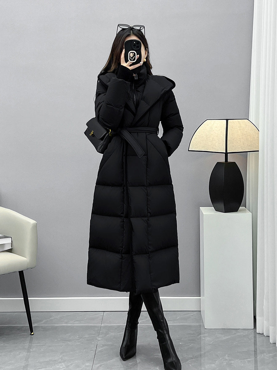 A full-length mirror selfie of the woman in the belted black puffer coat and tall boots, standing in a minimalist room with a modern floor lamp.