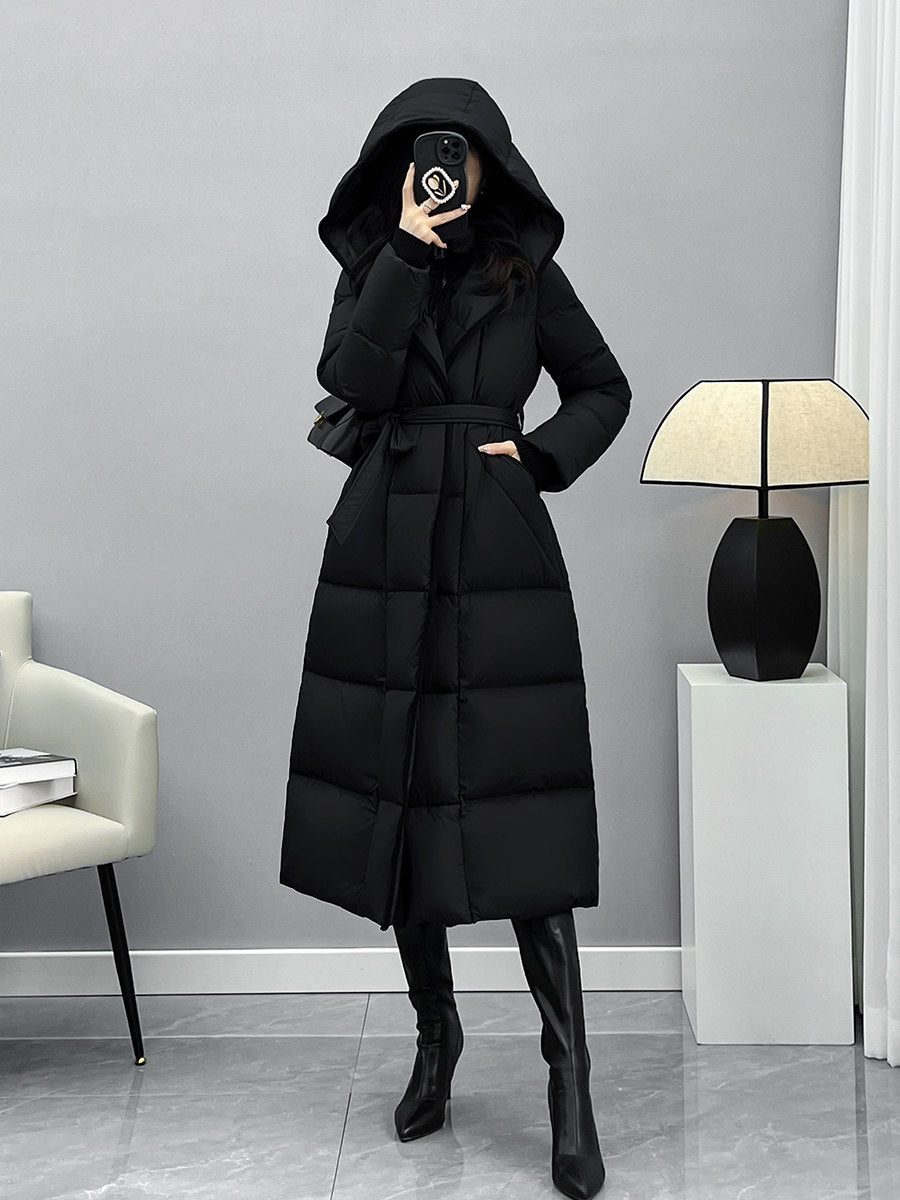 A full-length photo of a woman wearing a long, hooded black puffer coat with a cinched waist belt. She is wearing black leggings and pointed-toe black leather boots. Her face is obscured by her phone.