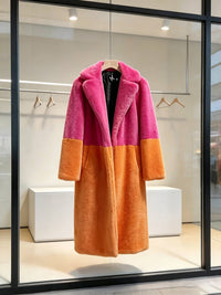 Bright pink and orange colorblock plush faux fur long coat displayed on a wooden hanger in a store window.