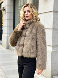 A woman wearing a cropped faux fur jacket with brown buckle details, standing outdoors in front of a stone building.