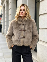 A woman wearing a cropped beige faux fur jacket with brown leather strap closures and black trousers, standing on a city sidewalk.