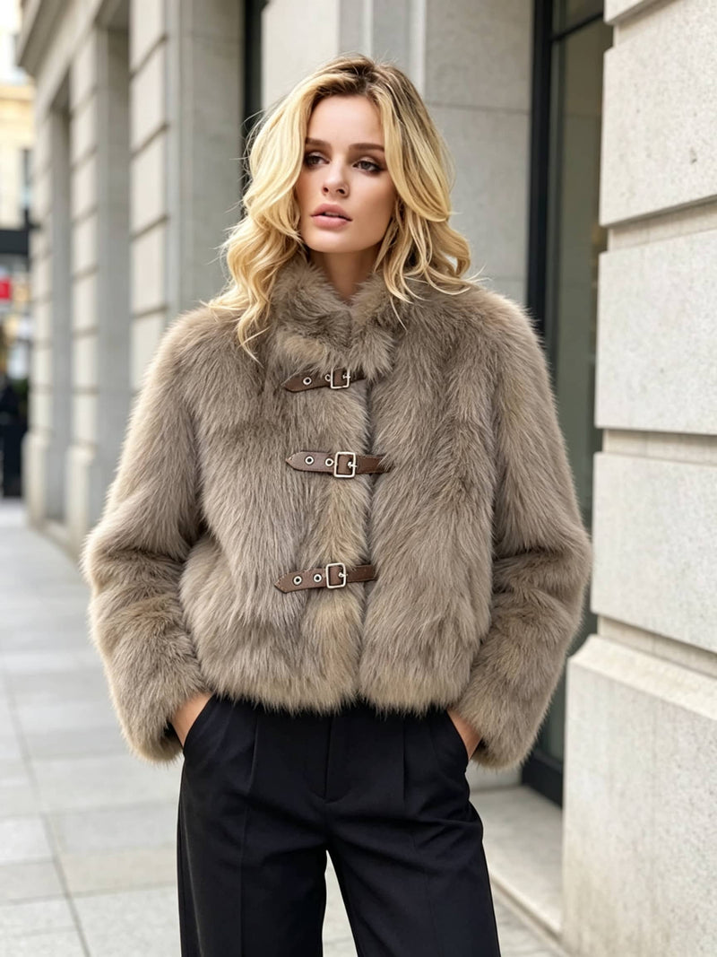 A woman wearing a cropped beige faux fur jacket with brown leather strap closures and black trousers, standing on a city sidewalk.