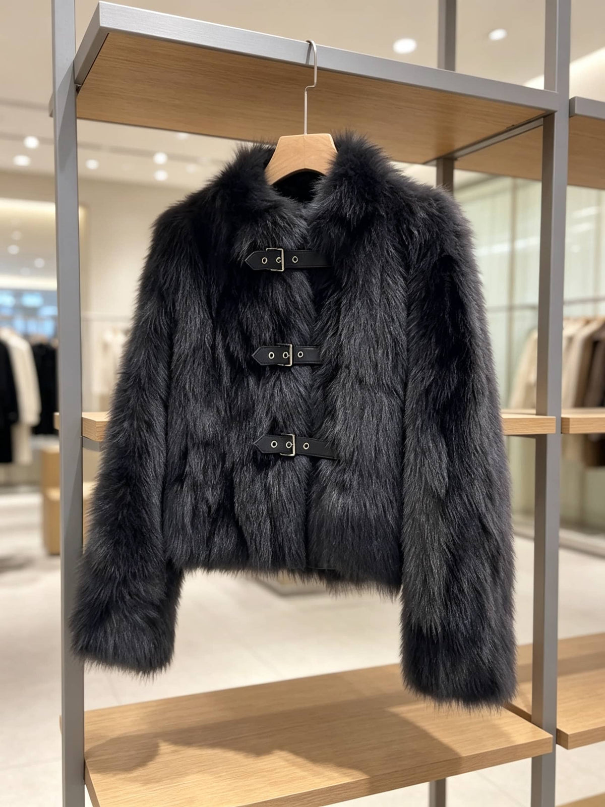 A dark gray faux fur jacket with black strap and silver buckle details, hanging on a wooden hanger on a clothing display rack inside a store.