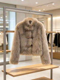 A light beige faux fur jacket with brown toggle closures, hanging on a wooden hanger from a metal clothing rack inside a boutique.
