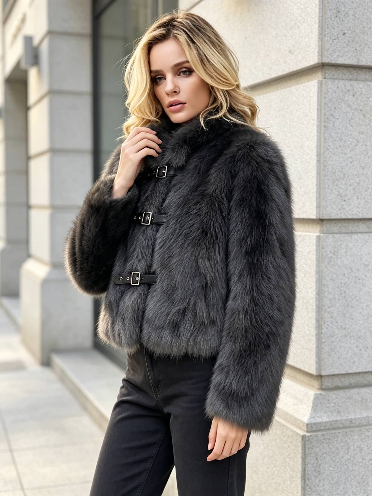 A woman wearing a dark gray faux fur jacket with black buckle straps, standing outdoors against a light gray stone building.