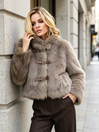 A woman wearing a light brown fur jacket with brown leather toggle closures and dark brown trousers, posing against a gray stone building on a city street.