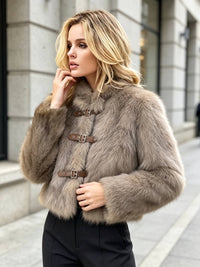 A woman wearing a plush taupe-brown faux fur jacket with brown leather and buckle toggle closures, standing outdoors on a city sidewalk.