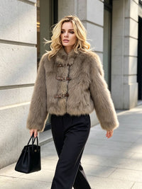 A woman walking outdoors in a beige faux fur cropped jacket with brown buckle details, paired with black trousers and holding a black handbag.