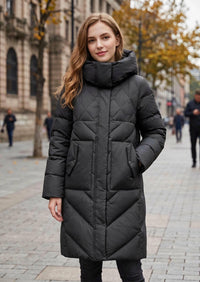 A woman wearing a long black quilted puffer coat with a high padded collar, standing on a city street with blurred buildings and trees in the background.