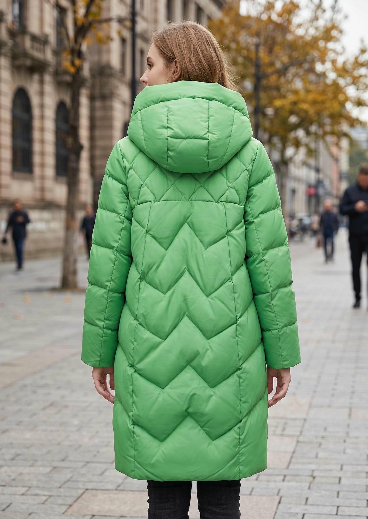 A woman viewed from behind wearing a bright green hooded puffer coat with a quilted chevron pattern, standing on a city street.