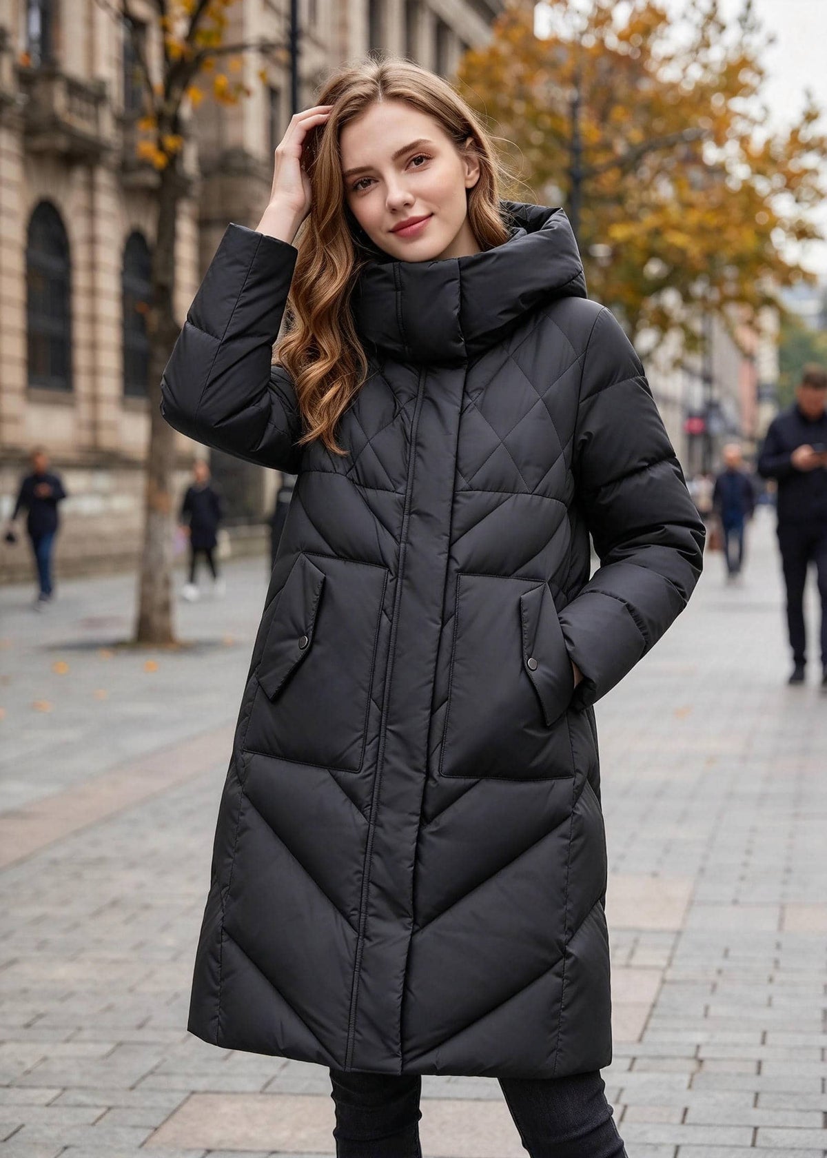 A woman wearing a long black quilted puffer coat with a high padded collar, standing on a city street with blurred buildings and trees in the background.
