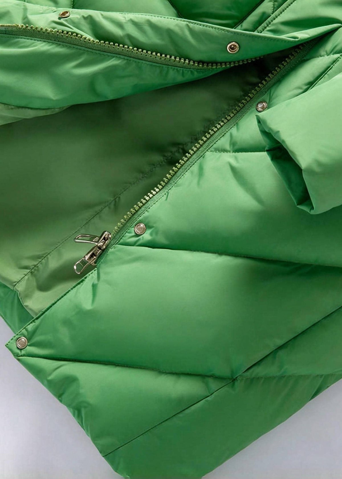 A close-up of a bright green puffer jacket showing the quilted stitching, a zipper closure, and metal snap buttons against a light background.