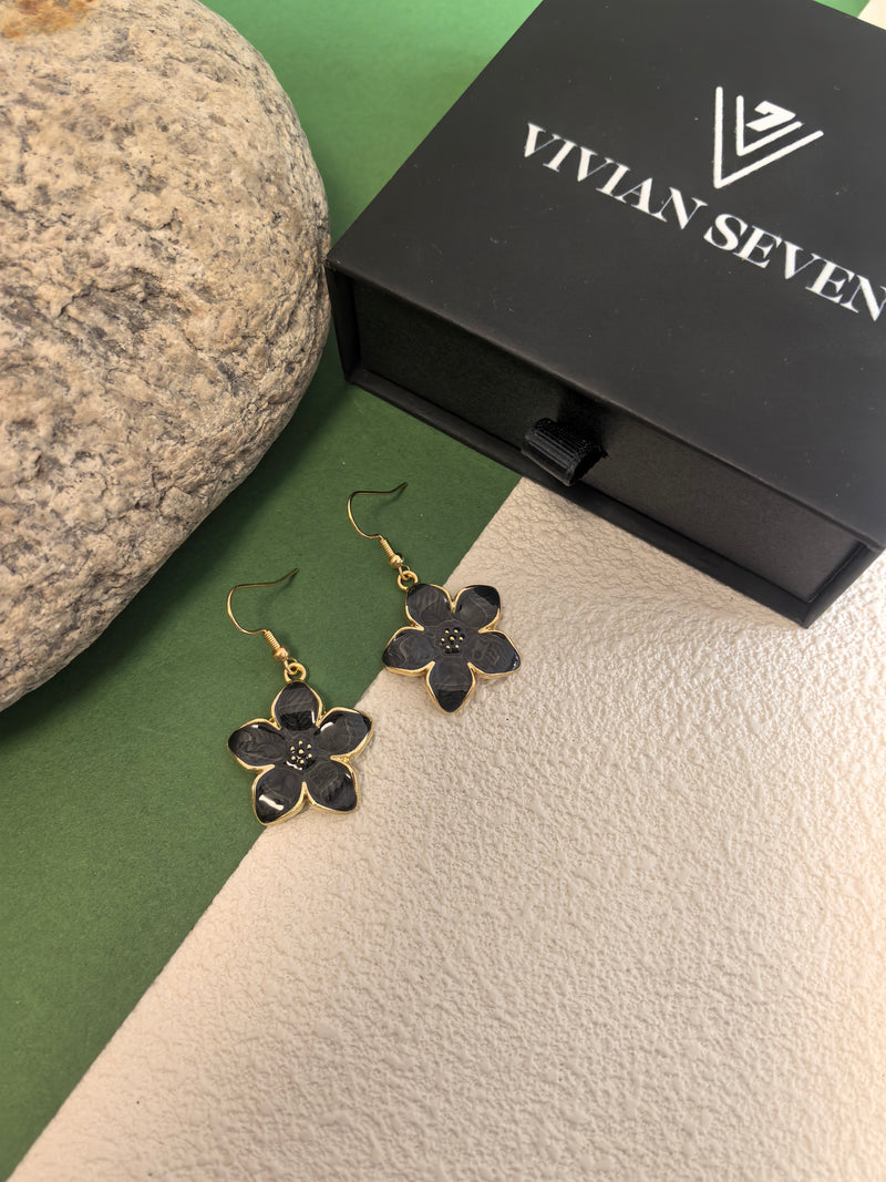 A pair of black enamel flower drop earrings with gold-toned trim, displayed outside of a closed black VIVIAN SEVEN branded box, resting on a background of green and textured beige.