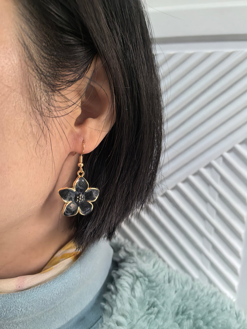 A close-up shot of a person wearing a grey enamel flower drop earring with a gold-toned fish hook wire in their earlobe.