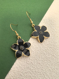 A close-up shot of a pair of black enamel flower drop earrings with gold-toned trim, showing the speckled detail in the center of the flowers, against a green and textured beige background.