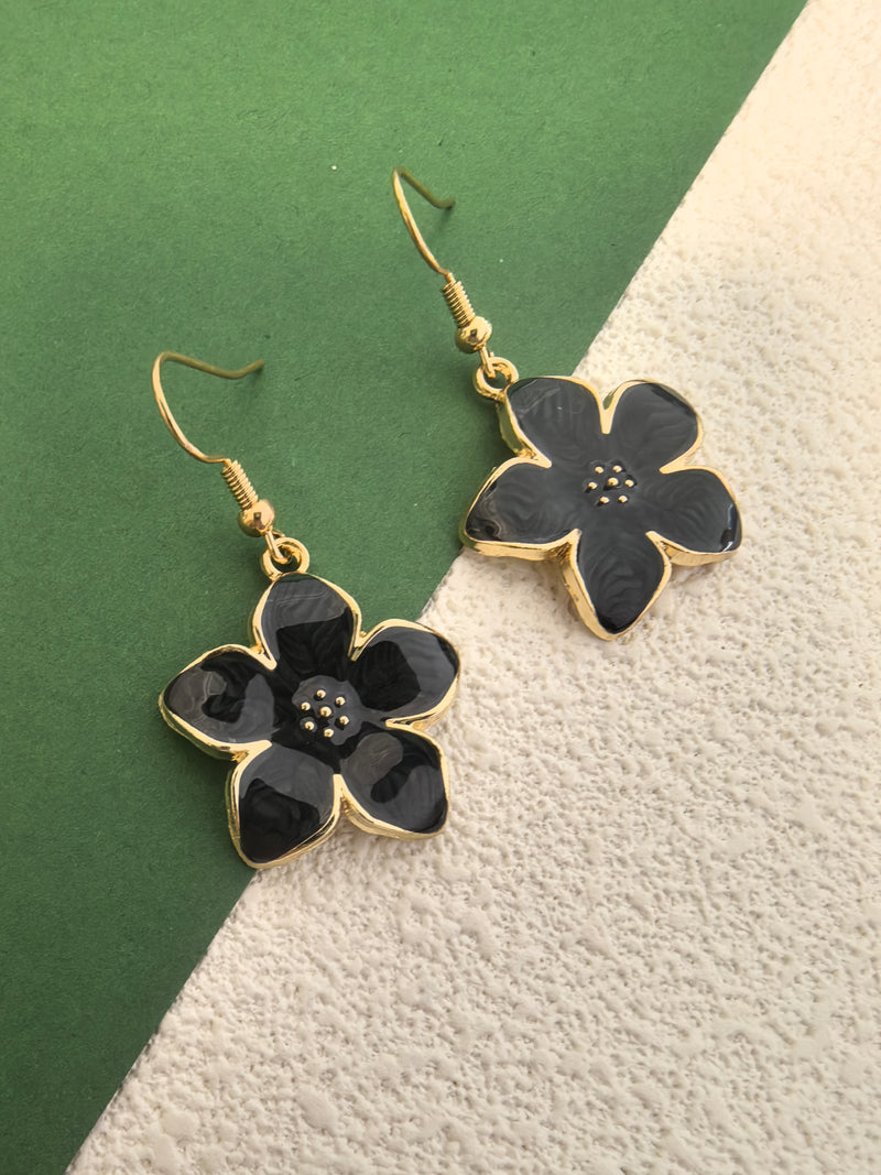 A close-up shot of a pair of black enamel flower drop earrings with gold-toned trim, showing the speckled detail in the center of the flowers, against a green and textured beige background.