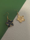 One black enamel flower drop earring with gold-toned trim placed next to another earring with only the gold-toned textured backing visible, against a green and textured beige background.