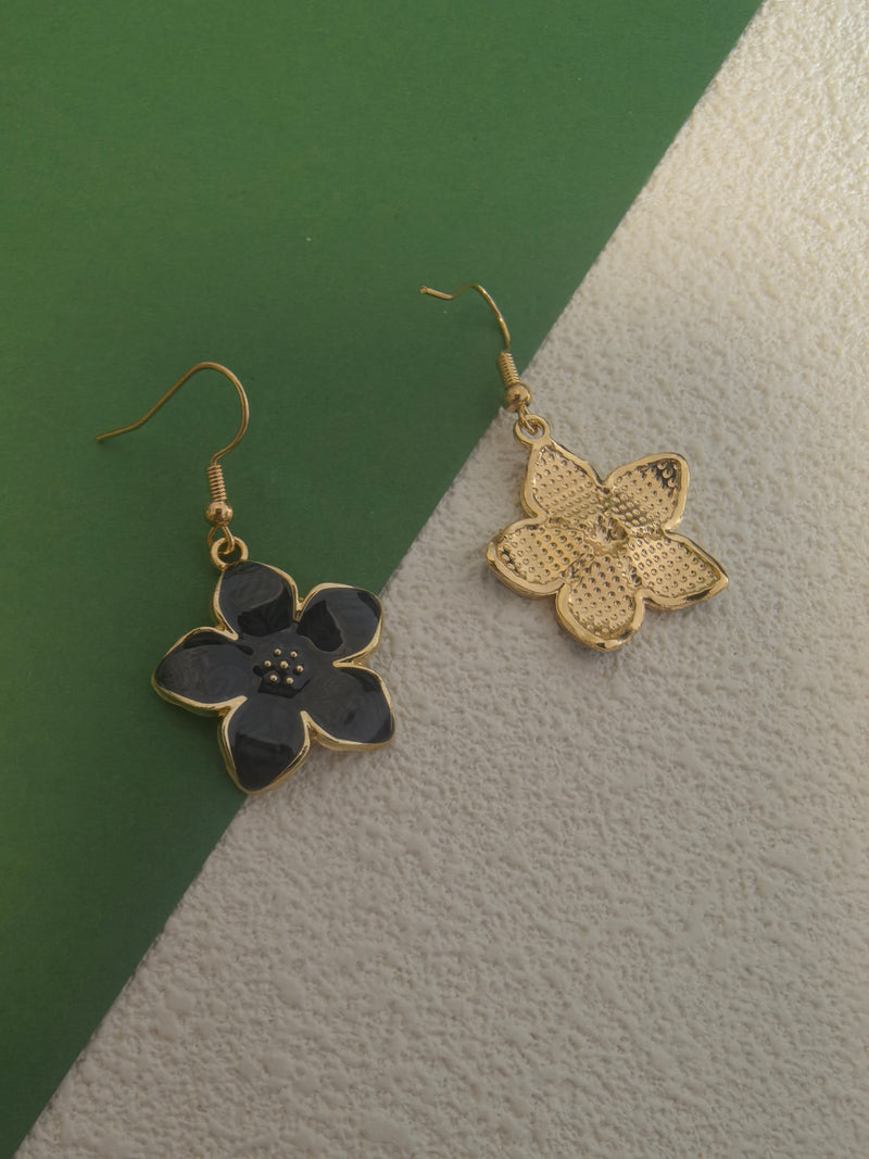 One black enamel flower drop earring with gold-toned trim placed next to another earring with only the gold-toned textured backing visible, against a green and textured beige background.