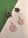 A collection of four enamel flower drop earrings: two black and two pink, all with gold-toned trim and fish hook wires, spread out on a background of green and textured beige.