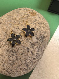 A pair of black enamel flower drop earrings with gold-toned trim and fish hook wires, resting on a textured large grey rock.