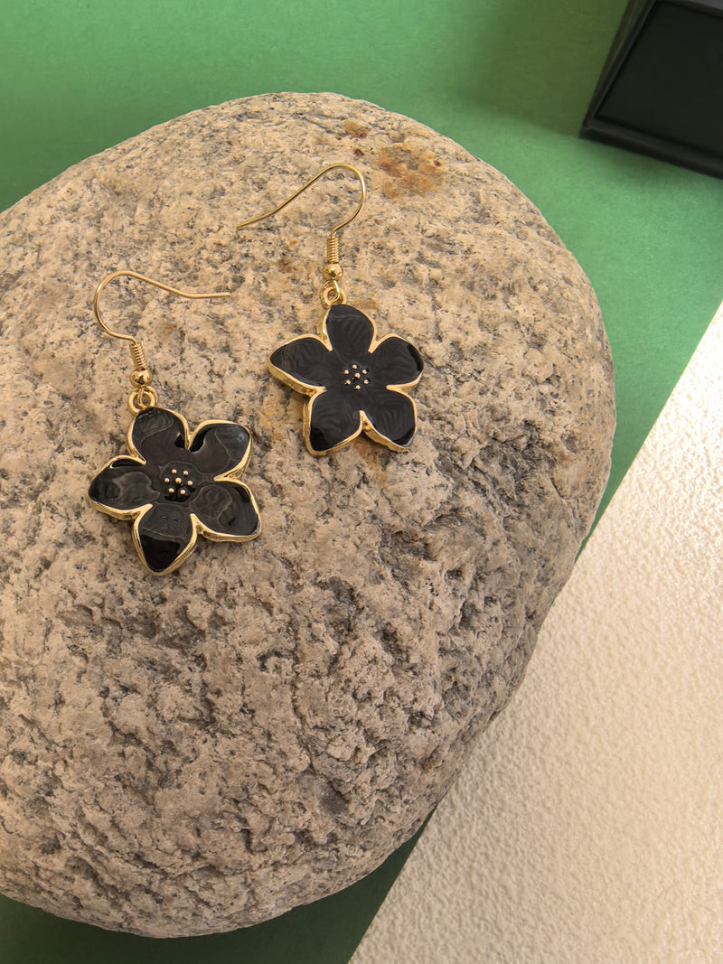 A pair of black enamel flower drop earrings with gold-toned trim and fish hook wires, resting on a textured large grey rock.