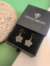 A pair of light grey enamel flower drop earrings with gold-toned trim and fish hook wires, displayed inside an open black VIVIAN SEVEN branded box on a black velvet inlay.