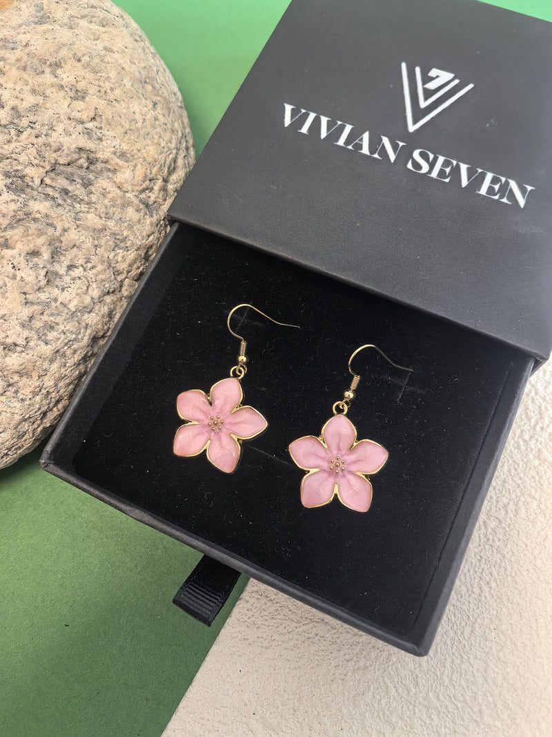 A pair of light pink enamel flower drop earrings with gold-toned trim and fish hook wires, displayed inside an open black VIVIAN SEVEN branded box on a black velvet inlay.