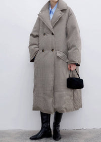 A person wearing a long beige and black houndstooth double-breasted coat over a light blue collared shirt, paired with black knee-high boots and holding a small black handbag against a plain light gray wall.