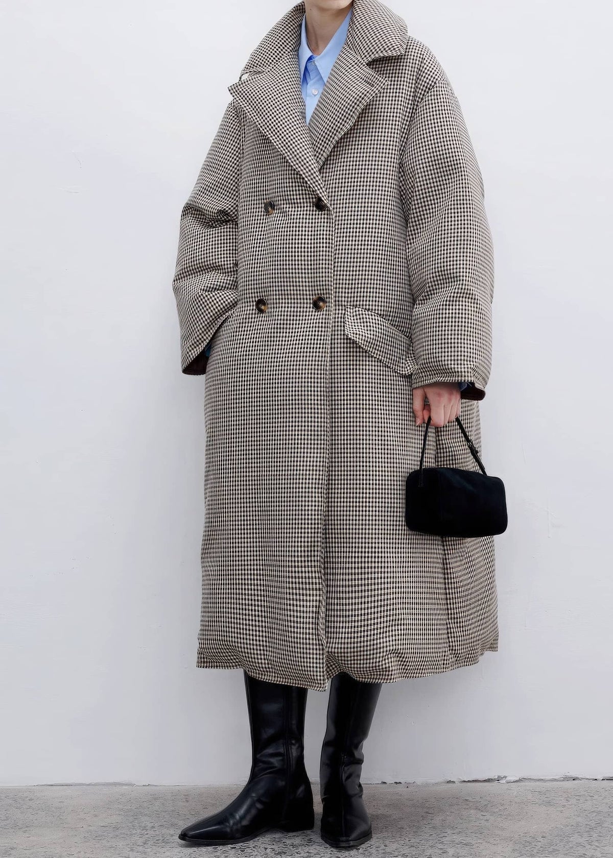 A person wearing a long beige and black houndstooth double-breasted coat over a light blue collared shirt, paired with black knee-high boots and holding a small black handbag against a plain light gray wall.