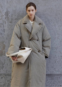 Three-quarter portrait of a woman wearing an oversized double-breasted plaid down maxi coat with wide lapels, holding an off-white leather shoulder bag against a concrete wall.