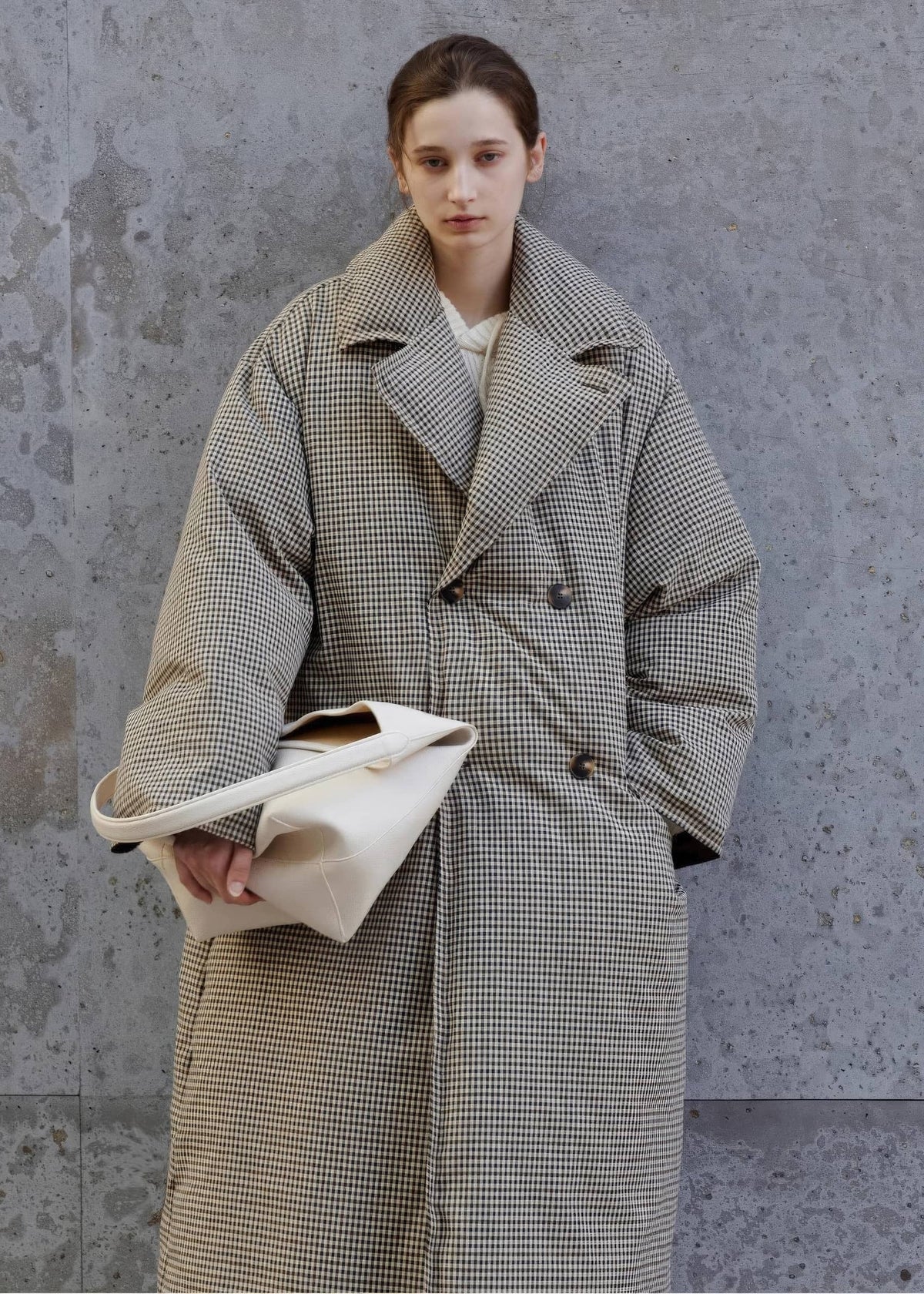 Three-quarter portrait of a woman wearing an oversized double-breasted plaid down maxi coat with wide lapels, holding an off-white leather shoulder bag against a concrete wall.