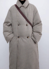 Cropped front view of a woman wearing a fully buttoned high-collar plaid down maxi coat with double-breasted tortoiseshell buttons and a crossbody strap across the chest.