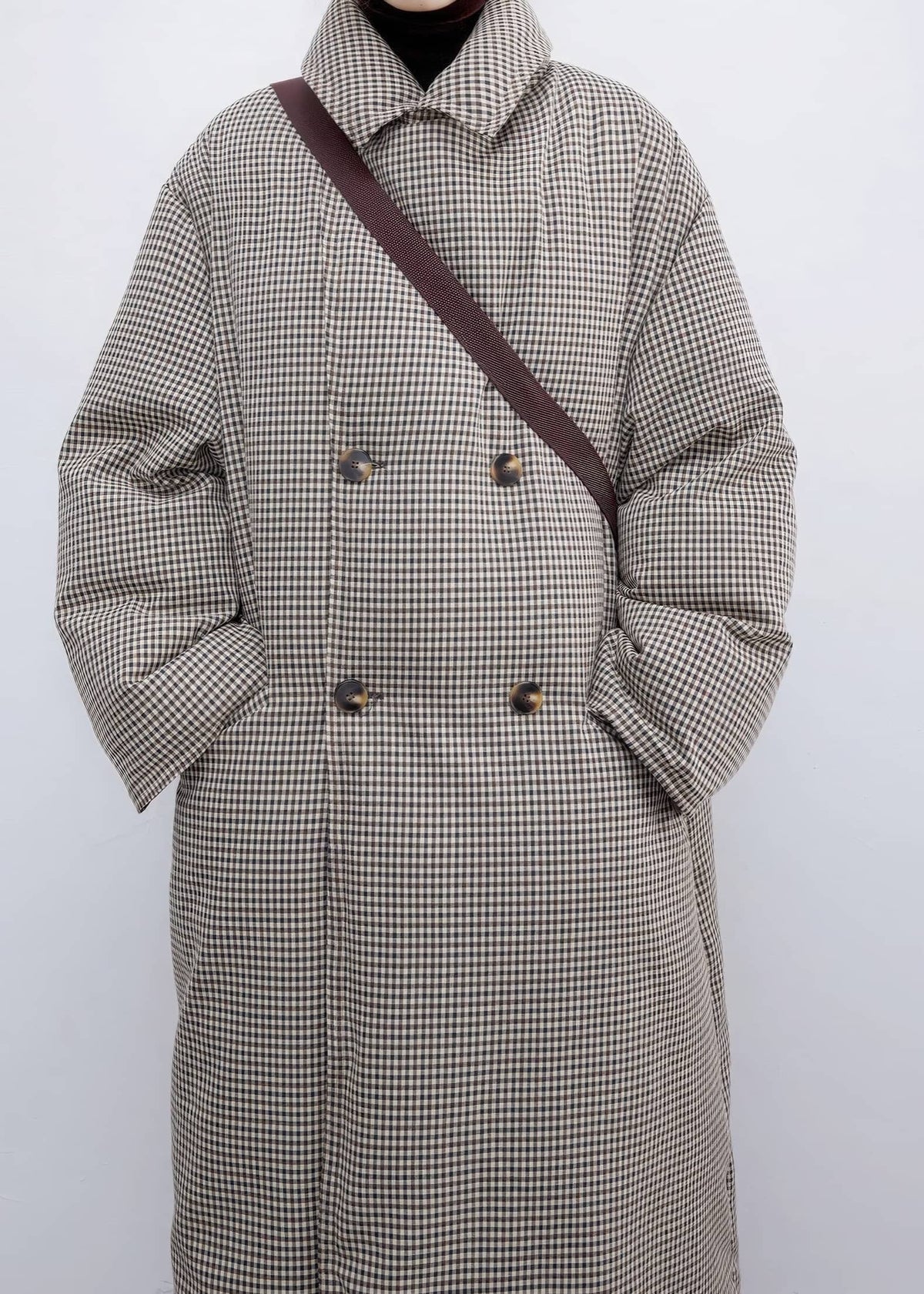 Cropped front view of a woman wearing a fully buttoned high-collar plaid down maxi coat with double-breasted tortoiseshell buttons and a crossbody strap across the chest.
