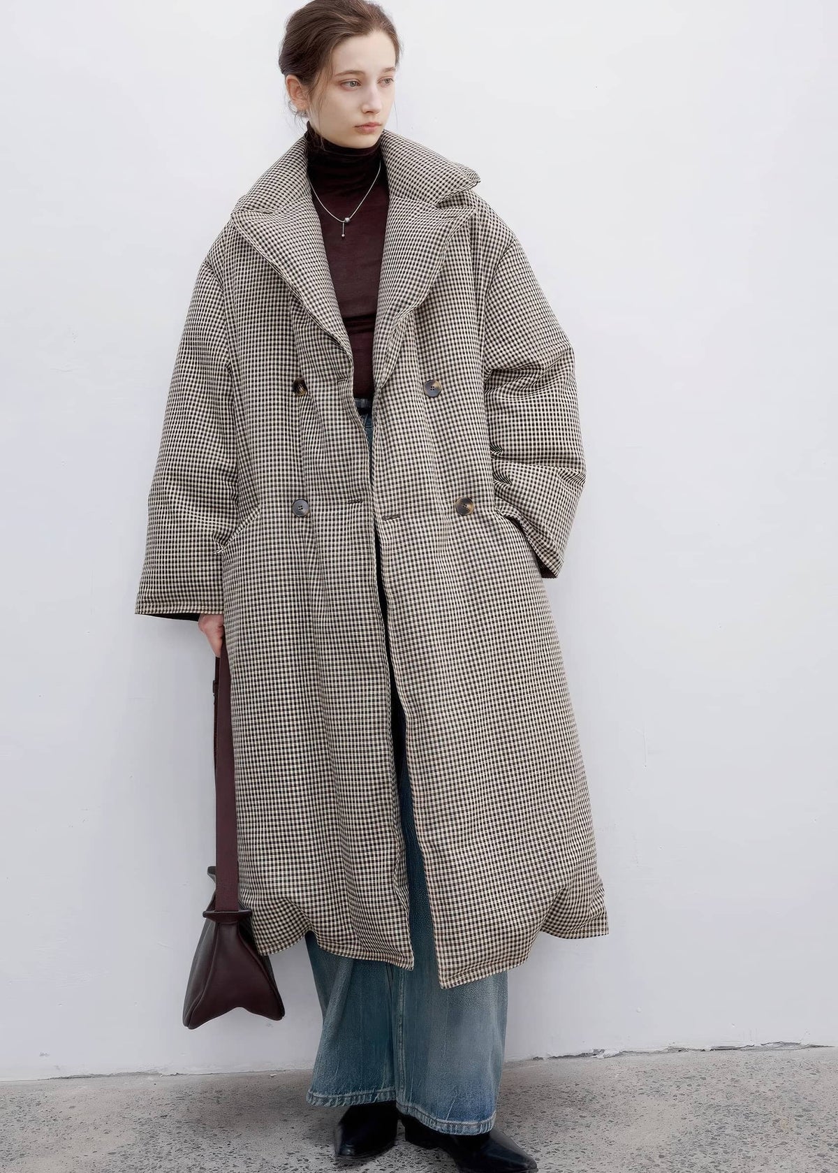 A model stands against a white wall wearing a long double-breasted houndstooth coat in beige and brown, layered over a dark brown turtleneck, blue jeans, and black shoes, while holding a dark brown shoulder bag.