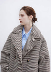 Profile close-up of a model wearing a double-breasted plaid down maxi coat with wide lapels over a light blue collared button-down shirt.
