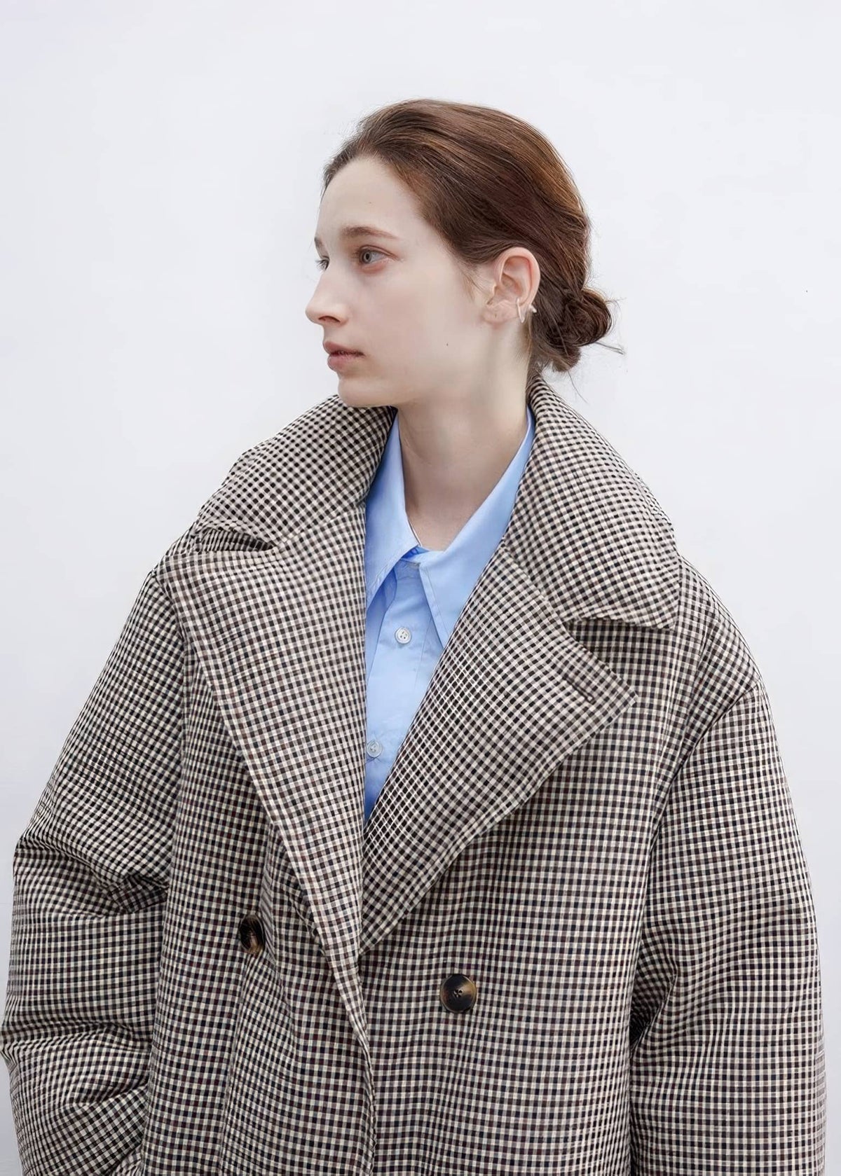 Profile close-up of a model wearing a double-breasted plaid down maxi coat with wide lapels over a light blue collared button-down shirt.