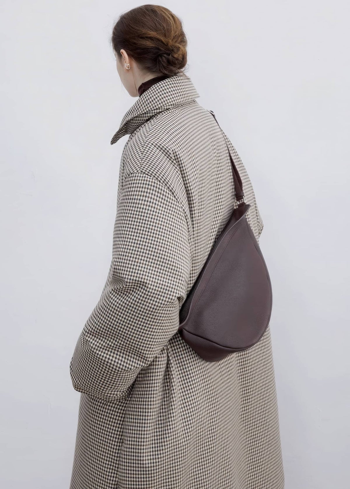 Back and side view of a model wearing a double-breasted plaid down maxi coat with a dark brown leather sling bag across the back.
