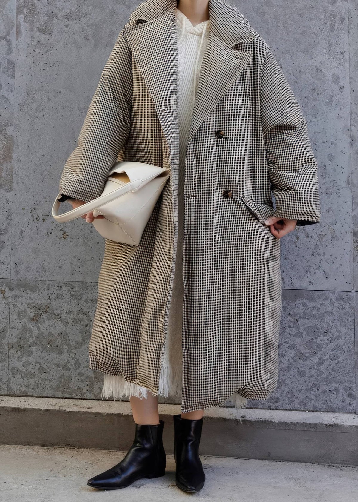 Cropped full-body street-style shot of a woman wearing an oversized plaid down maxi coat over a knit dress and carrying an off-white handbag.