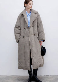 Full-length portrait of a model wearing a double-breasted plaid down maxi coat over a blue button-down shirt, finished with tall black leather boots.