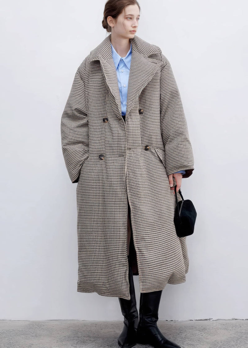 Full-length portrait of a model wearing a double-breasted plaid down maxi coat over a blue button-down shirt, finished with tall black leather boots.