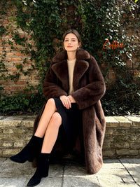 Woman sitting outdoors wearing a cozy dark coffee brown long faux fur coat with black ankle boots.
