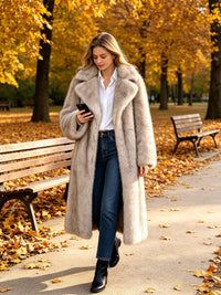  Woman looking at her phone while walking in an autumn park wearing a long light grey faux fur coat, white shirt, and jeans.