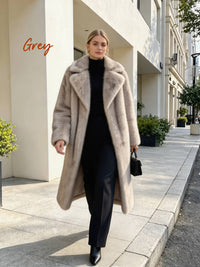 Full-length view of a model walking down a sidewalk in an open long grey faux fur coat carrying a black handbag.
