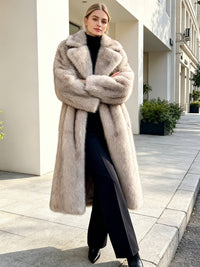 Woman wearing a long light grey faux fur coat with crossed arms, black turtleneck, and wide-leg trousers outdoors.