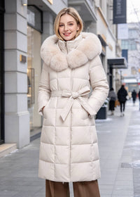 A woman wearing a long cream-colored puffer coat with a large fluffy faux-fur collar and a matching tie belt, standing on a city sidewalk.